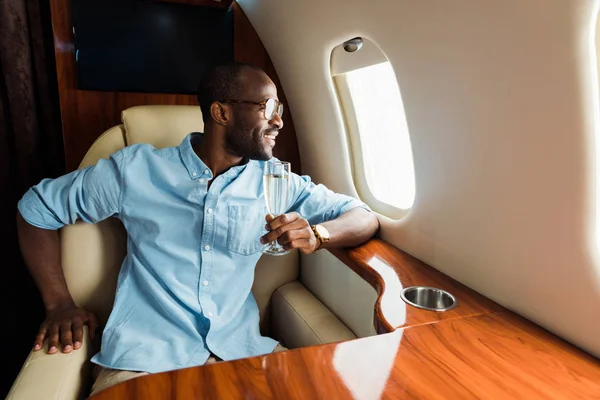 Allegro uomo africano americano in bicchieri che tengono bicchiere di champagne e guardando il finestrino dell'aereo — Foto stock