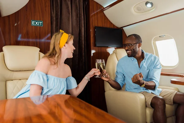 Selective focus of happy interracial couple clinking champagne glasses in private plane — Stock Photo