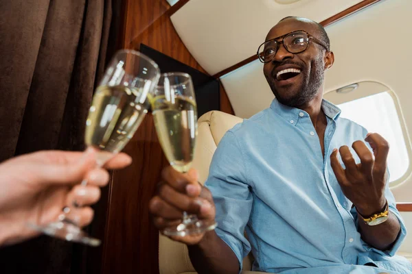 Selective focus of happy interracial couple clinking champagne glasses in private plane — Stock Photo