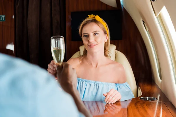 Selective focus of cheerful woman clinking champagne glasses with african american man in private plane — Stock Photo