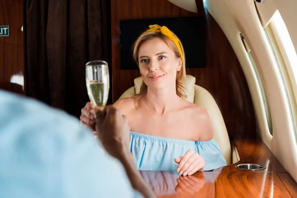 Messa a fuoco selettiva di donna allegra clinking bicchieri di champagne con l'uomo africano americano in aereo privato — Foto stock