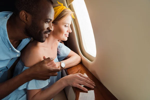 Hombre afroamericano feliz y mujer alegre mirando la ventana del avión en avión privado - foto de stock