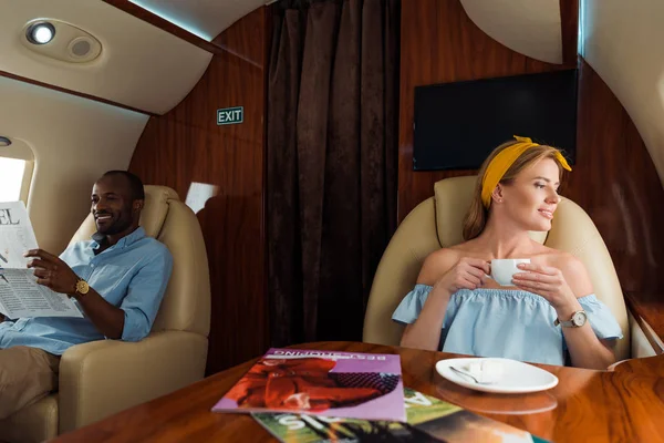 Fröhliche Frau hält Tasse mit Getränk in der Nähe eines afrikanisch-amerikanischen Mannes im Flugzeug — Stockfoto