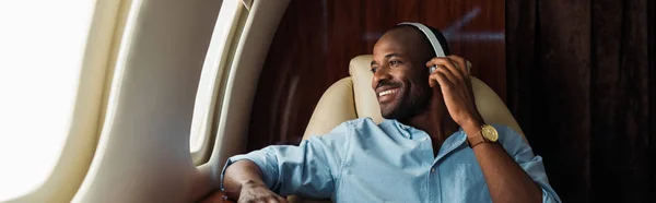 Panoramic shot of happy african american man listening music in private plane — Stock Photo