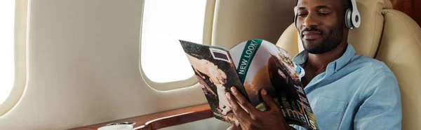 Tiro panorâmico do homem americano africano em fones de ouvido sem fio leitura revista em avião privado — Fotografia de Stock