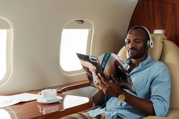 Selective focus of african american man in wireless headphones reading magazine in private plane — Stock Photo