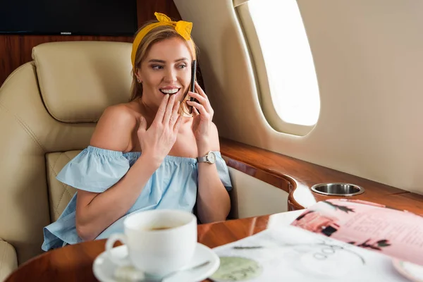 Selektiver Fokus einer glücklichen Frau, die mit einem Getränk im Flugzeug auf dem Smartphone in der Nähe der Tasse spricht — Stockfoto