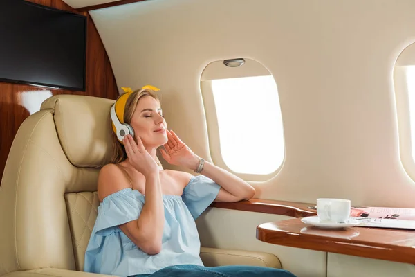 Mujer feliz escuchando música cerca de la taza con bebida en avión privado - foto de stock