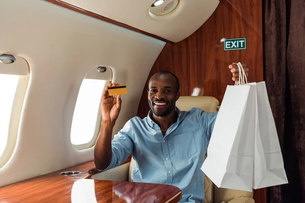 Happy african american man holding credit card and shopping bags in private plane — Stock Photo