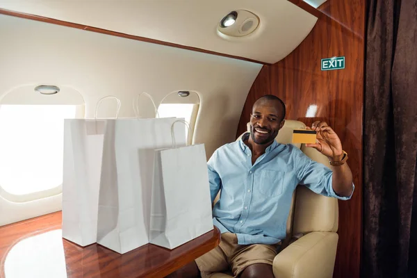 Alegre afroamericano hombre sosteniendo tarjeta de crédito y bolsas de compras en avión privado - foto de stock