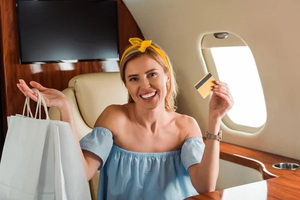 Alegre chica sosteniendo tarjeta de crédito y bolsas de compras en avión privado - foto de stock