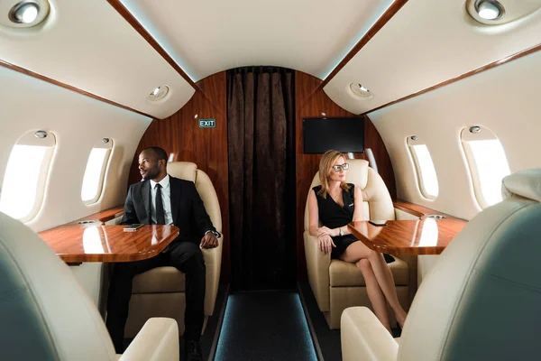 Selective focus of handsome african american businessman and attractive businesswoman sitting in private plane — Stock Photo