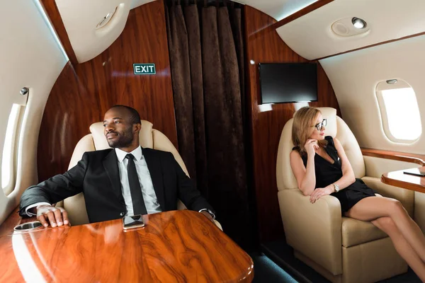 Selective focus of handsome african american businessman and beautiful businesswoman sitting in private plane — Stock Photo