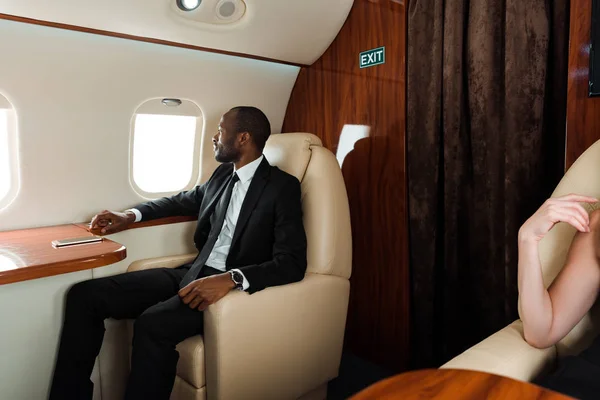 Apuesto hombre de negocios afroamericano mirando a la ventana del avión en avión privado - foto de stock
