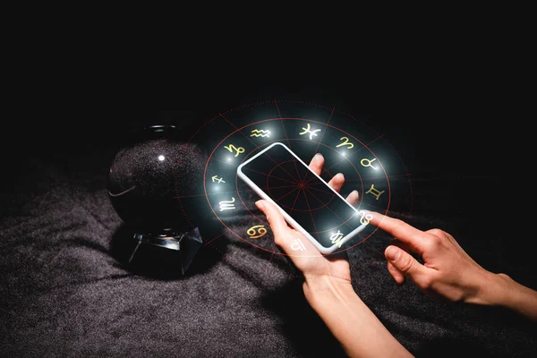 Ausgeschnittene Ansicht eines Astrologen, der ein Smartphone mit Tierkreiszeichen in der Nähe der Kristallkugel auf schwarzem Samtstoff hält — Stockfoto