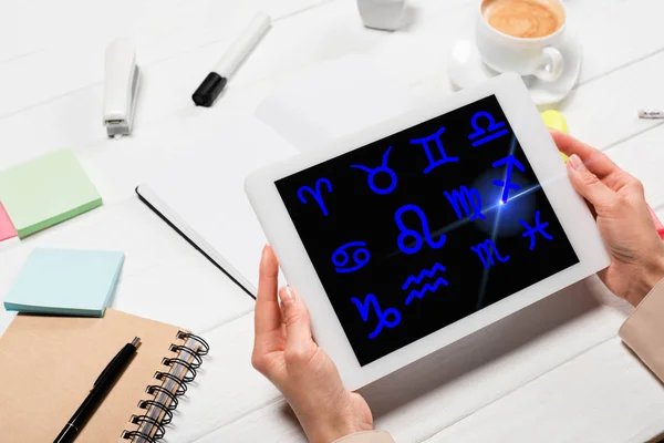 Vue recadrée de la femme tenant tablette numérique avec signes du zodiaque sur le lieu de travail avec fournitures de bureau et café — Photo de stock