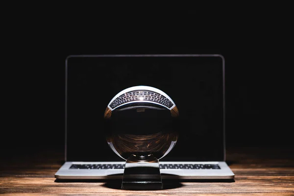 Bola de cristal cerca del ordenador portátil en la mesa de madera sobre fondo negro - foto de stock