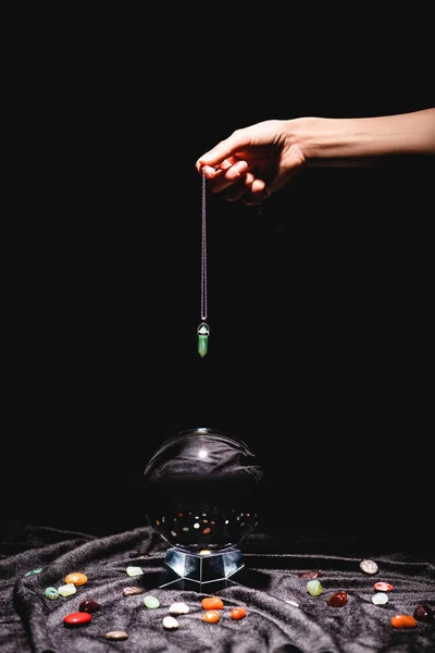 Cropped view of fortuneteller holding stone above crystal ball with fortune telling stones on black velvet cloth isolated on black — Stock Photo