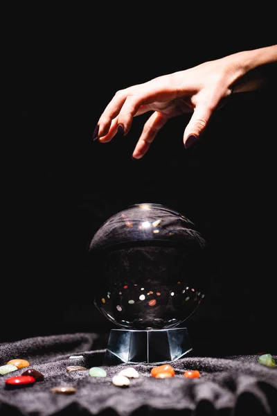 Cropped view of fortuneteller hand above crystal ball with fortune telling stones on black velvet cloth isolated on black — Stock Photo