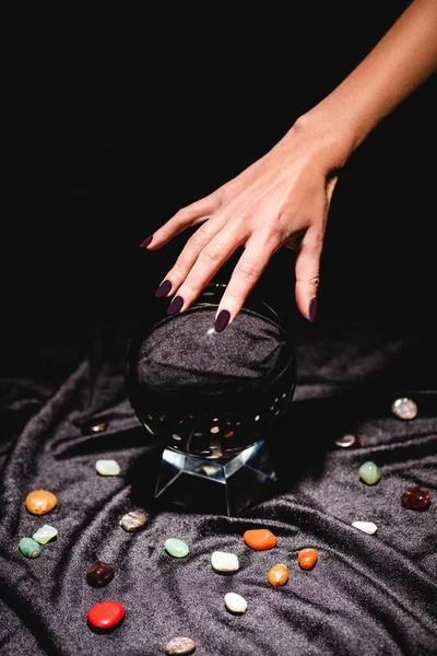 Cropped view of fortuneteller hand above crystal ball with fortune telling stones on black velvet cloth isolated on black — Stock Photo