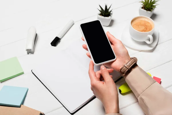 Vista recortada de la mujer sosteniendo teléfono inteligente en el lugar de trabajo con suministros de oficina y café - foto de stock