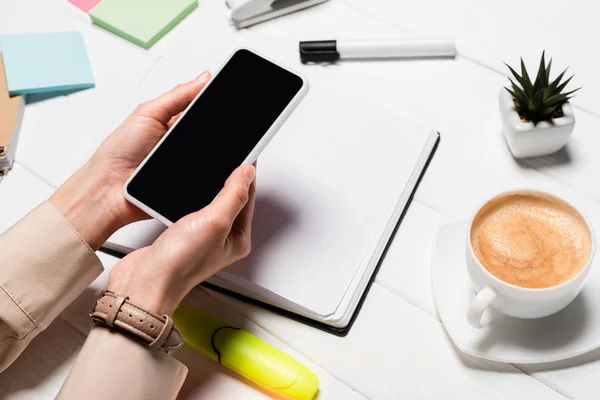 Cropped view of woman holding smartphone at workplace with office supplies and coffee — Stock Photo