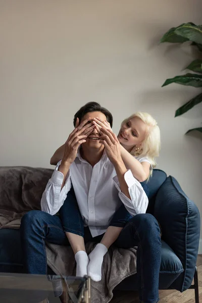 Sonriente joven que cubre los ojos del novio con las manos en el sofá en la sala de estar - foto de stock