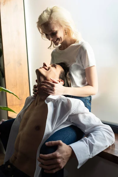 Sonriente chica tocando sin camisa novio mientras está sentado en alféizar de la ventana - foto de stock