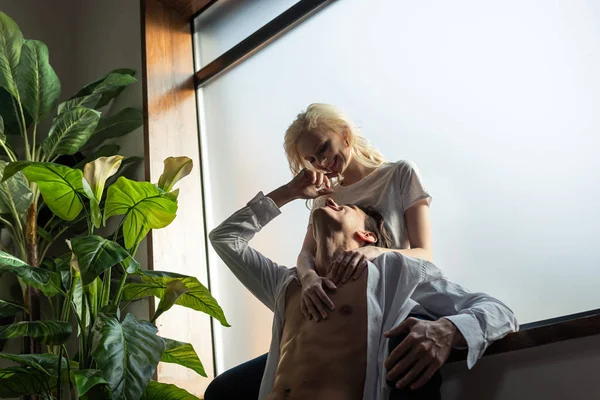 Улыбающаяся женщина обнимает мужчину без рубашки у окна в гостиной — стоковое фото
