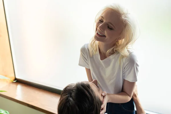 Sourire femme blonde toucher petit ami visage wile assis sur le rebord de la fenêtre — Photo de stock