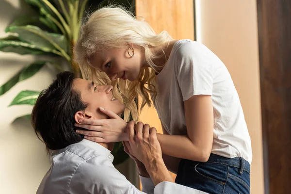 Vue latérale d'une fille blonde souriante touchant le visage de son petit ami à la maison — Photo de stock