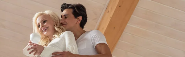 Низкий угол зрения улыбающийся мужчина обнимает подругу с кофе дома, панорамный снимок — стоковое фото