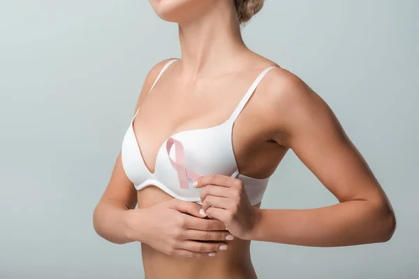 Vista recortada de la mujer joven en sujetador blanco con cinta rosa como conciencia de cáncer de mama aislado en gris - foto de stock