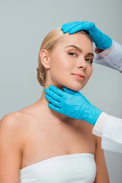 Vista recortada de esteticista en guantes de látex azul tocando mujer aislada en gris - foto de stock