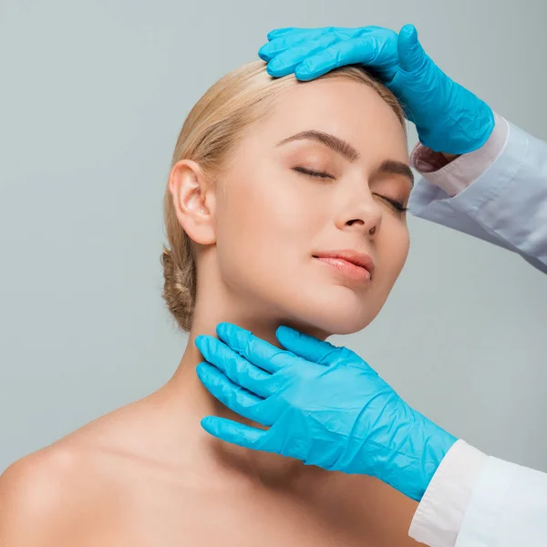 Abgeschnittene Ansicht einer Kosmetikerin in blauen Latex-Handschuhen, die nackte Frau mit geschlossenen Augen berührt, isoliert auf grau — Stockfoto
