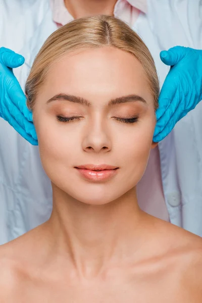 Cropped view of beautician in blue latex gloves touching temples of woman with closed eyes — Stock Photo
