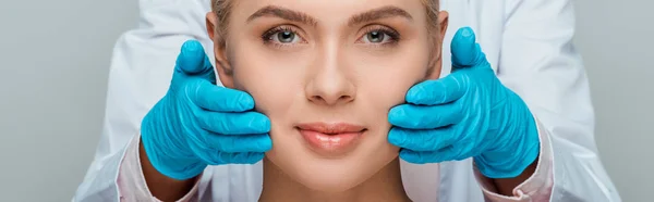 Панорамный снимок косметолога в голубых латексных перчатках касающихся щек женщины, изолированной на сером — стоковое фото