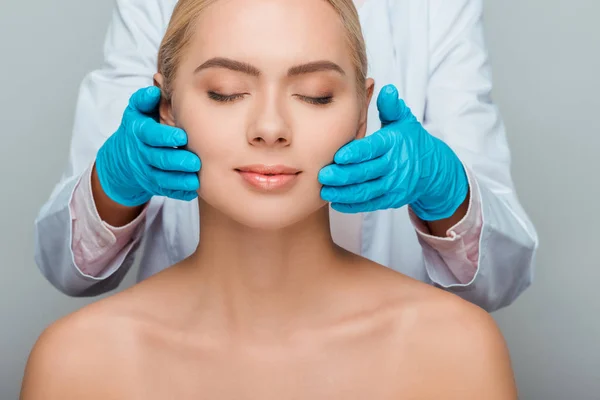 Cropped view of beautician in blue latex gloves touching cheeks of naked woman isolated on grey — Stock Photo