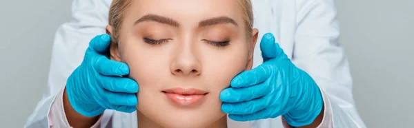 Prise de vue panoramique de l'esthéticienne en gants de latex bleu touchant les joues de la femme avec les yeux fermés isolés sur gris — Photo de stock