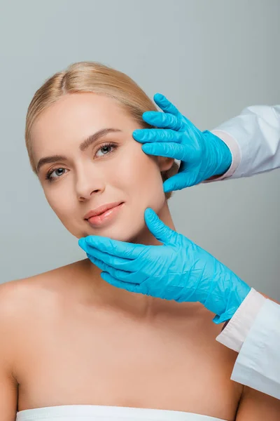 Vista recortada de esteticista en guantes de látex azul tocando la cara de mujer joven aislada en gris - foto de stock