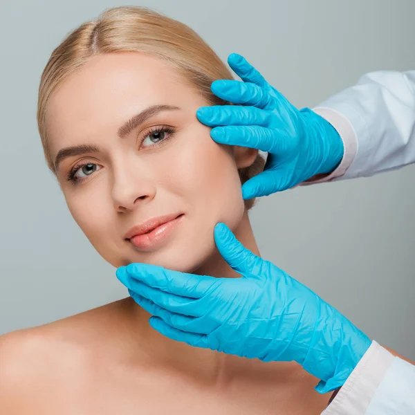 Abgeschnittene Ansicht der Kosmetikerin in Latex-Handschuhen berührt Gesicht der jungen Frau isoliert auf grau — Stockfoto