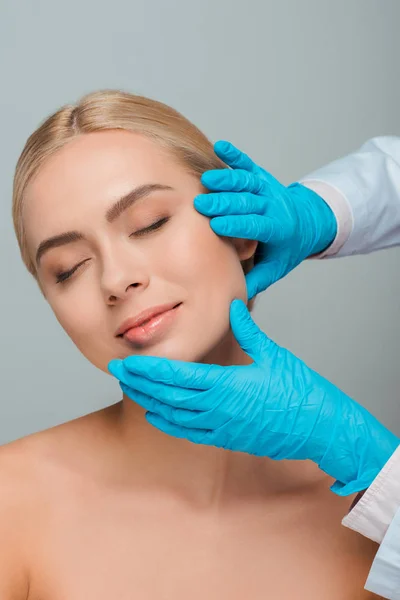 Vista recortada de esteticista en guantes de látex tocando la cara de la joven feliz aislado en gris - foto de stock