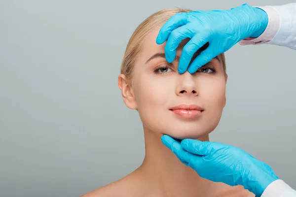 Vista recortada de esteticista en guantes de látex tocando la nariz de mujer desnuda feliz aislado en gris - foto de stock