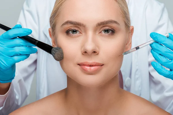 Cropped view of beautician in latex gloves holding cosmetic brush and syringe near attractive girl — Stock Photo