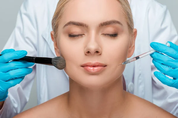Cropped view of beautician in latex gloves holding cosmetic brush and syringe near attractive girl with closed eyes — Stock Photo
