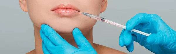 Panoramic shot of beautician in latex gloves holding syringe near lips of woman isolated on grey — Stock Photo