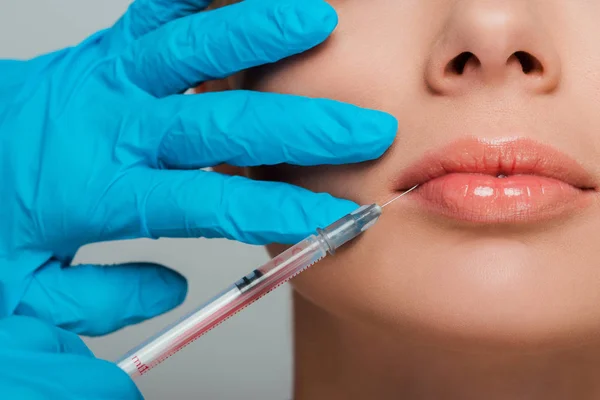 Vista recortada de esteticista en guantes de látex con jeringa cerca de los labios de chica aislada en gris - foto de stock