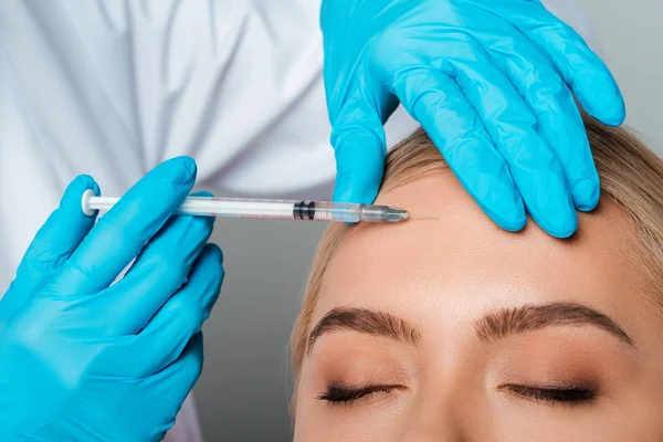 Vista cortada de esteticista em luvas de látex segurando seringa perto da testa da menina isolada em cinza — Fotografia de Stock