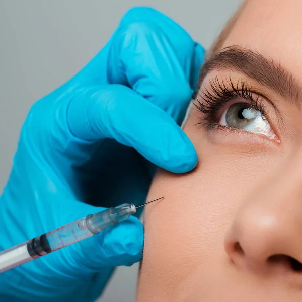 Primer plano de esteticista en guante de látex sosteniendo jeringa cerca de mejilla de chica aislada en gris - foto de stock