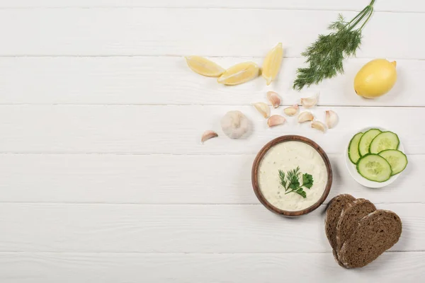 Top view of tzatziki sauce with ingredients and bread on white wooden background — Stock Photo